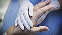 Bildet viser et par hender med plasthansker som holder hånda til en gammel person