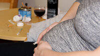 Bildet viser magen til en gravid dame som sitter i en stol. I hånda holder hun en sigarett. På bordet bak står et askebeger fullt av sneiper og et glass med rødvin. Noen pilleglass ligger veltet på bordet.