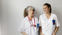 Bildet viser en ung og en eldre sykepleier som smiler til hverandre.