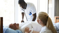 The photo shows an older patient lying in a hospital bed. A male nurse is bending towards him listening with a stethoscope. A female nurse is sitting beside the bed.