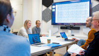 The photo shows two students sitting around a table opposite two academic supervisors. A third student can be seen with her back towards the camera. At the screen on the wall graphs of cardiac arrhythmias can be seen. The people are participating in a reflection group.