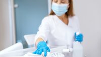 Bildet viser en sykepleier med munnbind og hansker. Hun holder en sprayflaske i hånden og tørker med en fille