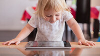 Bildet viser en smilende, liten jente som står og kikker ned på er nettbrett som ligger på bordet