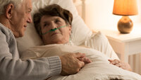 Bildet viser en syk eldre dame som ligger i senga med neseslange. En eldre mann holder henne i hånda og kikker på henne