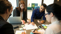 Bildet viser gruppearbeid på et kontor, der flere mennesker sitter rundt et bord med PC, ark og gule lapper foran seg.