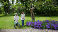 Bildet viser en eldre dame med rullator som er på tur i en hage med en yngre dame.
