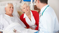 Bildet viser en eldre mann som ligger i senga på sykehuset. Kona sitter ved siden av, og legen er på visitt.