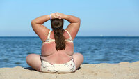 Bildet viser en overvektig kvinne bakfra, som sitter på stranden i bikini.