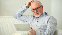 Bildet viser en eldre mann som sitter i en sofa med et nettbrett på fanget og klør seg litt i hodet