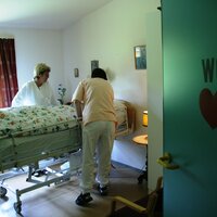 Bildet viser en pasient i en seng på sykehjem, med to pleiere som står ved sengen.