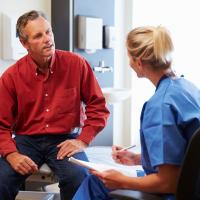 Bildet viser en mannlig pasient som er i konsultasjon med en kvinnelig sykepleier. Han sitter på en sykehusseng, og hun har et skjema i hånda.