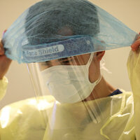 Bildet viser en mann som tar på seg beskyttelsesutstyr, som maske, visir, smittefrakk osv.