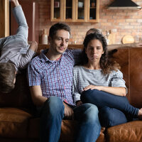 Bildet viser en mann og en kvinne som ser slitne ut og sitter i en sofa, og et barn som klatrer på sofaryggen