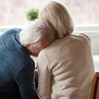 Bildet viser et eldre par som sitter inntil et bord. Mannen lener hodet sitt på kvinnens skulder.