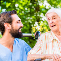 Bildet viser en mannlig sykepleier som snakker med en eldre dame. De sitter ute, og det er god stemning.