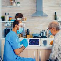 Bildet viser en mannlig sykepleier i blå uniform, munnbind og visir på hjemmebesøk hos en eldre mann, også han med munnbind. De sitter og ser på en ipad.