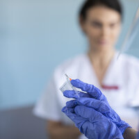 Bildet viser en hånd som holder en sprøyte, og en kvinne i bakgrunnen.