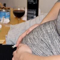 Bildet viser magen til en gravid dame som sitter i en stol. I hånda holder hun en sigarett. På bordet bak står et askebeger fullt av sneiper og et glass med rødvin
