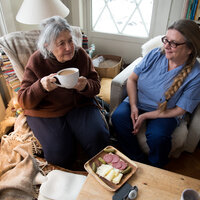 Bildet viser en beboer som drikker kaffe sammen med hjemmesykepleieren. Foran beboeren står et fat med påsmurte brødskiver.